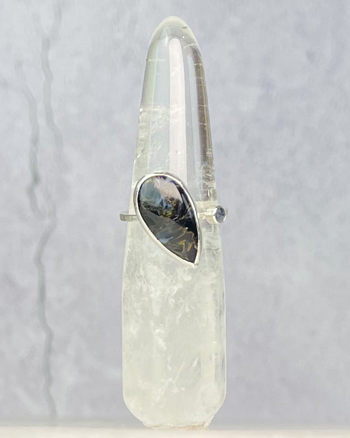 A clear quartz crystal point tower with a silver-toned ring holding a teardrop-shaped iridescent black and gold stone, set against a textured light gray background.